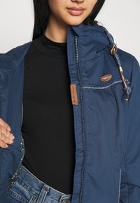 Mörkblå lättviktsjacka med huva, dragkedjestängning och elastiska detaljer, har en brun läderlogotyp och ett mönstrat foder.