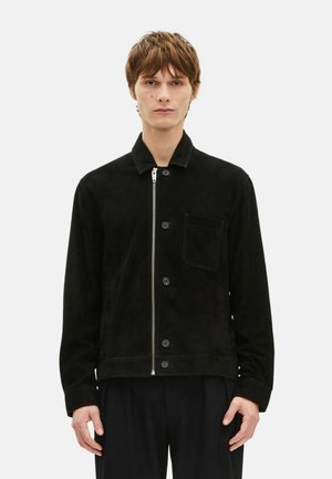 Młody mężczyzna w czarnej koszuli i kurtce na zamek oraz guziki z kieszenią na piersi, stoi na tle jednolitego białego tła.