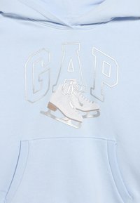 Φωτεινό γαλάζιο φούτερ με ασημένια γραμμή "GAP" και λευκά γραφικά παγοπέδιλα πάνω από την εμπρόσθια τσέπη.