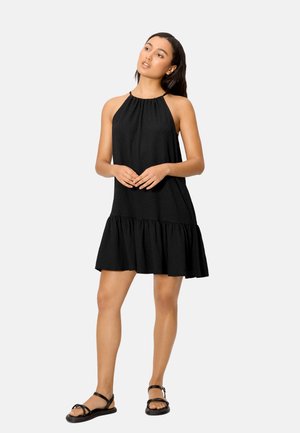 Mujer de pie vistiendo un vestido negro sin mangas con cuello halter, dobladillo con volantes y sandalias planas negras sobre un fondo liso.