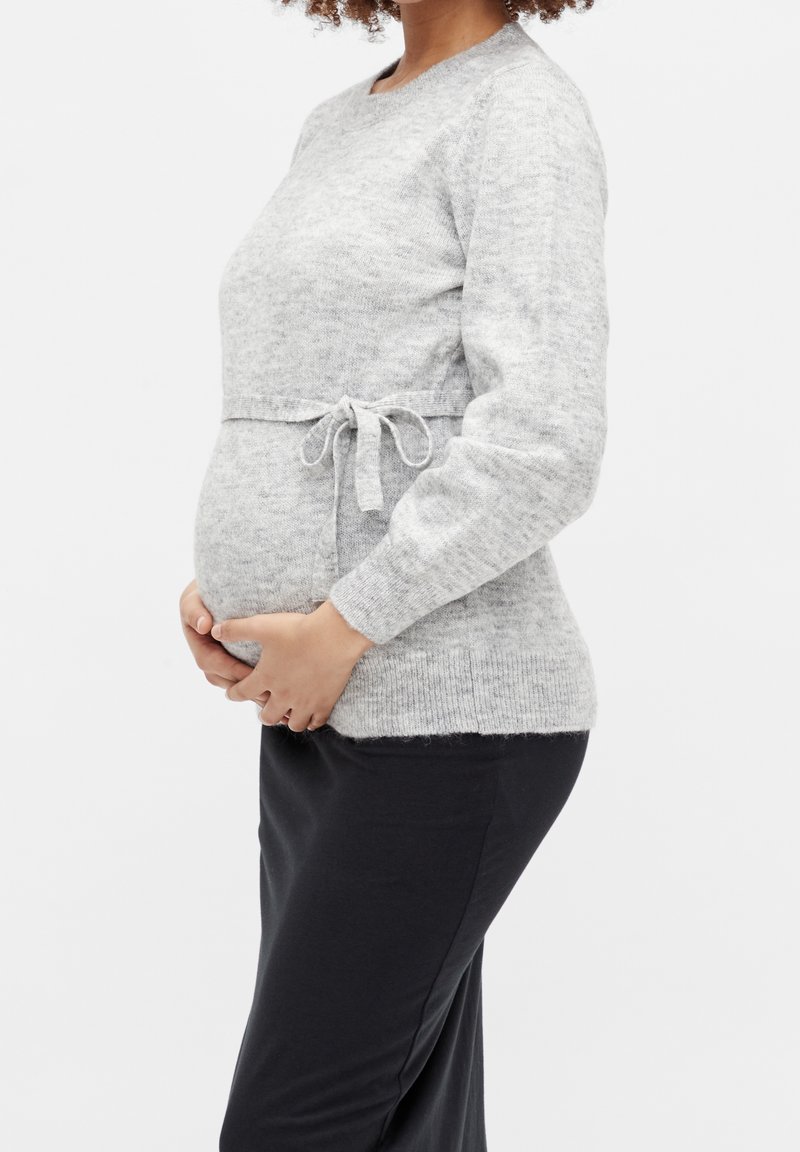 Femme enceinte portant un pull tricoté gris clair avec une ceinture nouée, tenant son ventre, debout devant un fond blanc uni.