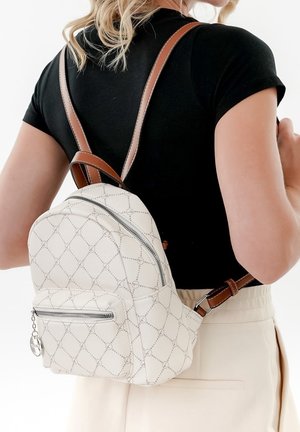 Kremowy plecak z wzorem w romby, brązowymi paskami, przednią kieszenią na zamek i srebrnymi okuciami. Materiał wydaje się gładki i trwały.