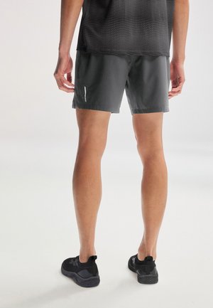 Set bagfra af en person iført sorte sportsshorts, sort nettop og sorte atletiksk sko, stående på en lys baggrund.