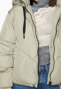 Gewatteerd jack in lichtgrijs, met een capuchon, zwarte ritssluiting en gestructureerde stof. Draagt over een top met "wolk"-patroon en blauwe spijkerbroek.