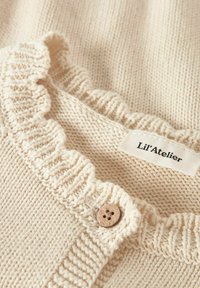 Beżowy sweter dzianinowy z falbaniastym kołnierzem, z drewnianym guzikiem i metką z napisem "Lil'Atelier." Teksturowany materiał z widocznymi stębnowaniami.