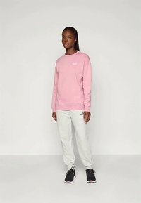 Sieviete stāv, ģērbusies rozā džemperī, gaiši pelēkās sporta biksēs un melnās sporta kurpēs uz vienkārša gaiša fona.