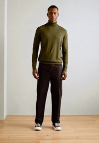 Maglione a collo alto verde oliva, pantaloni cargo neri e sneakers bianche e nere. Tessuto morbido, design aderente e tasche pratiche.