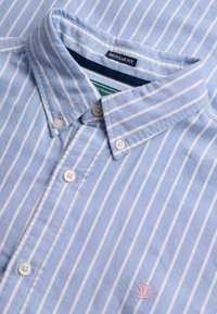 Hellblaues Hemd mit Knöpfen und weißen vertikalen Streifen, bezeichnet als „Regular Fit“, mit kleinem rosa gesticktem Logo nahe dem Saum.