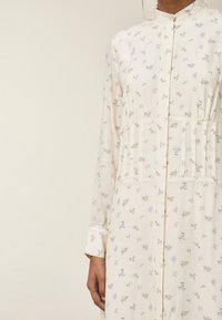 Långärmad klänning i krämfärg med små blå och bruna blommönster, med hög krage och vertikala paneldetaljer vid midjan.