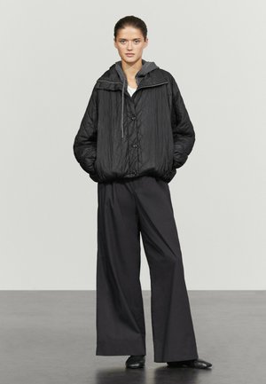 Frau steht und trägt eine schwarze gesteppte Jacke mit einem grauen Kapuzenpullover darunter und weite schwarze Hosen.