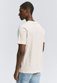 Tricou ivory cu mânecă scurtă, realizat din bumbac moale, având un guler clasic în formă de cerc și un mic detaliu cu etichetă pe lateral.