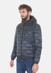 Giacca a vento imbottita grigio scuro con cappuccio, caratterizzata da una trama trapuntata e tasca sul petto con cerniera. Indossata sopra jeans in denim.