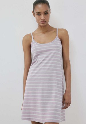 Žena u svijetloljubičastoj haljini bez rukava s bijelim horizontalnim prugama stoji ispred jednobojne svijetlosive pozadine.