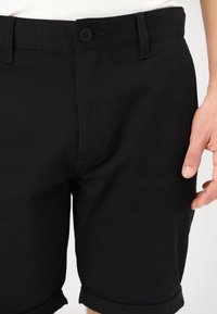 Shorts en coton noir avec fermeture à bouton, devant plat et revers roulés. Texture lisse avec des détails de couture subtils.