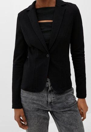 Osoba nosząca dopasowaną czarną marynarkę z jednym guzikiem na czarnym topie z poziomymi wycięciami, w połączeniu z szarymi, wysokim stanem, jeansami z efektem sprania.