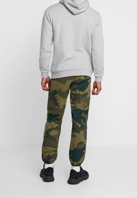 Hombre vestido con sudadera gris, pantalones de camuflaje verde y azul oscuro, calcetines blancos y zapatillas negras, de pie contra un fondo claro y liso.
