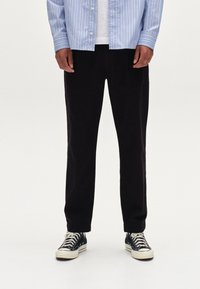 Pantalon noir à coupe droite, porté avec une chemise rayée bleu clair et des baskets noires. Le tissu semble lisse avec une texture minimale.