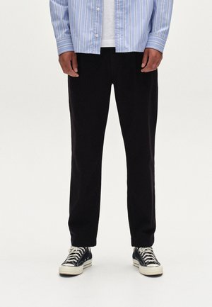 Schwarze Hosen mit geradem Schnitt, getragen mit einem hellblauen gestreiften Hemd und schwarzen Sneakers. Der Stoff wirkt glatt mit minimaler Textur.