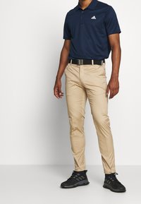 Muž stojí, oblečený v námořnickém polo tričku, béžových kalhotách a černých sportovních botách, s jednoduchým bílým pozadím za ním.