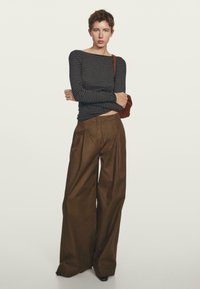 Persoon met kort haar in een zwart en grijs gestreepte lange mouw shirt, brede bruine broek en een bruine schoudertas.