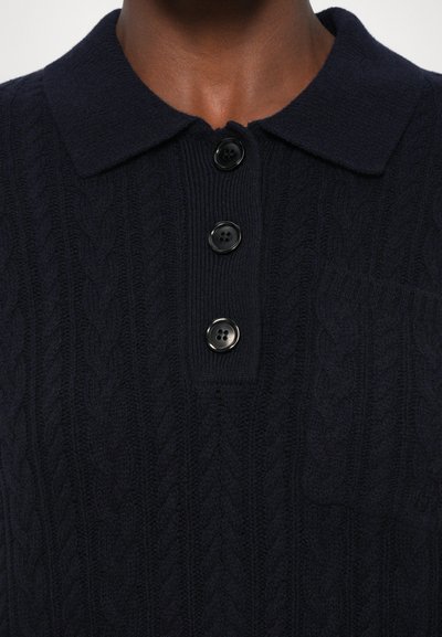 Marineblå kabelstrikket polo med krage, med fire svarte knapper og en brystlomme. Den teksturerte stoffet gir dybde til designet.