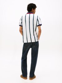 Weißes Poloshirt mit blauen und grünen vertikalen Streifen, marinblauem Kragen und roten Akzenten. Kombiniert mit dunkelblauen Jeans und braunen Schuhen.
