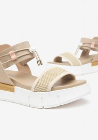 Beige platform sandals with a textured mesh strap, elastic band, and white chunky sole. Features a decorative hardware accent and logo.