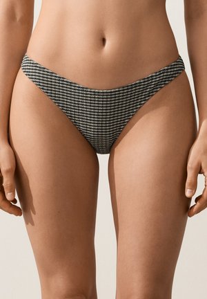 Femeie care poartă slip de bikini cu talie înaltă și model în carouri alb-negru, stând pe un fundal simplu, deschis la culoare.