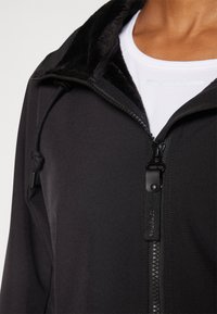 Veste noire zippée avec un col doux et texturé, dotée d'une fermeture éclair et d'un cordon de serrage réglable. Tissu lisse et design ajusté.