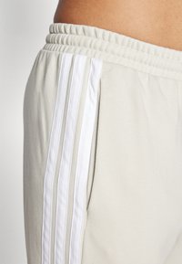 Šviesiai smėlinės kelnės su raukšlėtu liemeniu, šoniniais kišenėmis ir trimis baltais vertikaliais dryžiais išoriniame siūle. Minkšta tekstūra.