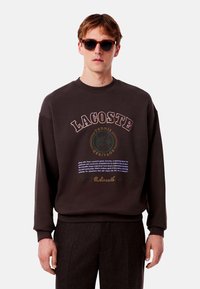 Sudadera marrón oscuro con un gran logo de Lacoste, detalles en texto y puños acanalados. El modelo lleva gafas de sol y pantalones a medida.