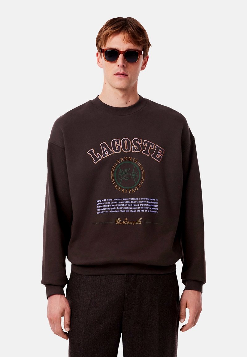 Ciemnobrązowy sweatshirt z dużym logo Lacoste, detalami tekstowymi i ściągaczami przy mankietach. Model nosi okulary przeciwsłoneczne i dopasowane spodnie.