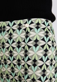 Gemusterte high-waisted Hose mit einem geometrischen Design in Pastellfarben und Schwarz, verziert mit einem metallischen Logo-Accente und einer glatten Stofftextur.