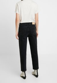 Jean noir taille haute à coupe droite, avec deux poches arrière et un patch en cuir marron sur la ceinture. Haut blanc et bottes à motifs visibles.