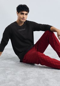 Cămașă neagră cu mânecă lungă cu logo-ul "Calvin Klein"; pantaloni de pijama roșii cu un model mic repetitiv. Așezat pe un covor gri deschis.