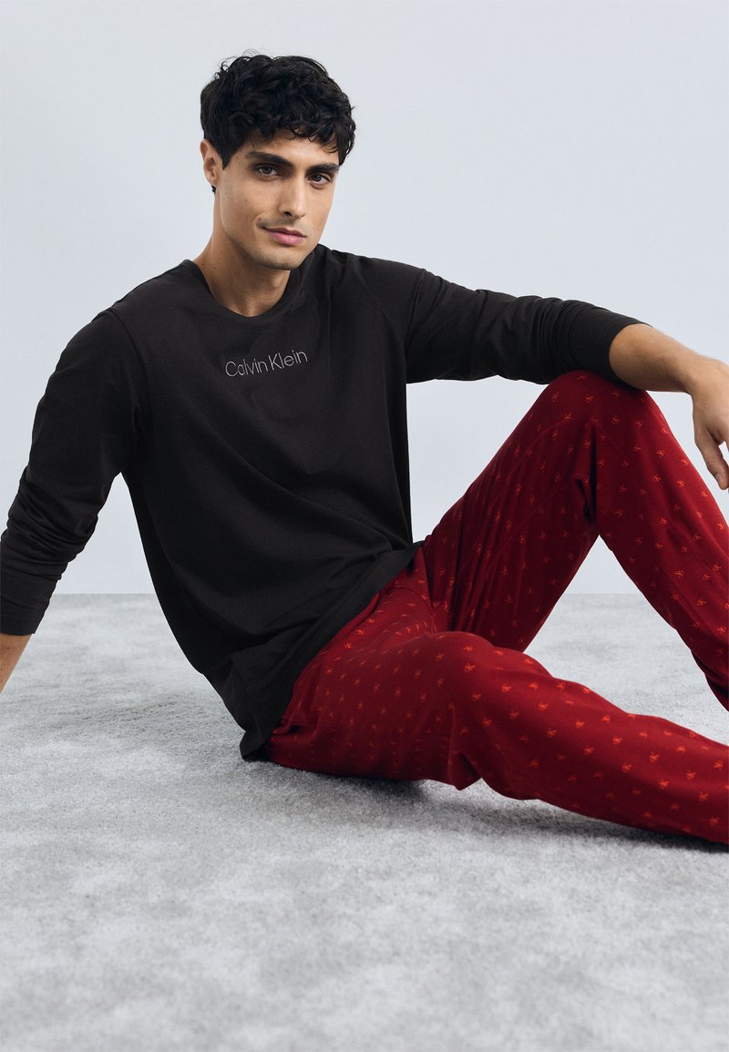 Cămașă neagră cu mânecă lungă cu logo-ul "Calvin Klein"; pantaloni de pijama roșii cu un model mic repetitiv. Așezat pe un covor gri deschis.