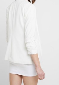 White tailored blazer with a structured fit, shoulder pads, and rolled-up sleeves. Pairing with a short white skirt. Smooth fabric texture.