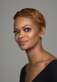 Korte haarsnit, warme bruin tint, gladde textuur. Zwart topje, lage halslijn. Parel oorbellen, goed gedefinieerde wenkbrauwen en zacht geblend make-up.