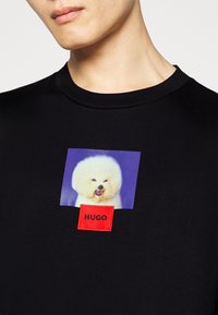 Sort kortærmet t-shirt med et grafik af en hvid fluffy hund og et rødt rektangulært mærke med "HUGO" trykt i sort.