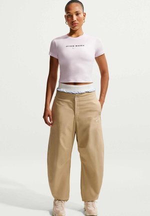 Vrouw staat met een lichtroze cropped Nike Shox t-shirt, beige wijde broek, witte tailleband en roze sneakers tegen een effen witte achtergrond.