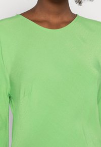 Top a maniche lunghe di un brillante verde, realizzato in un tessuto liscio e strutturato. Presenta un ampio scollo e un design minimalista senza motivi o dettagli visibili.