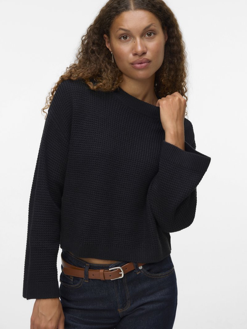 Zwarte gebreide trui met een ontspannen pasvorm, brede mouwen en een cropped ontwerp. Gepaard met donkerdenim jeans en een bruine riem.