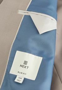 Švarkas slim fit stiliaus, pagamintas iš kreminės ir šviesiai mėlynos medžiagos, su balta etikete, nurodančia "NEXT", dydis 40R, ir lygiu pamušalo paviršiumi.