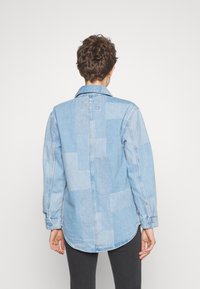 Světle modrá denimová košile s patchworkovým designem, dlouhými rukávy, límečkem a zaobleným dolním okrajem. Viditelné švy a manžety na knoflíky.