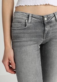 Los jeans de mezclilla gris presentan un diseño ajustado con bolsillos estándar y un cierre de botón. La textura es suave con una ligera apariencia desgastada.