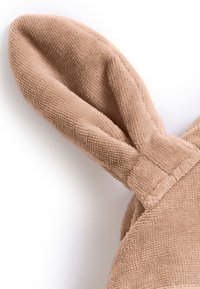 Beige tyg med en texturerad, ribbad yta. Har mjuka, vikta kaninöron fästa vid en rundad bas. Ingen synlig hårdvara.