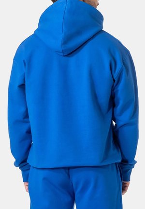 Hombre con sudadera con capucha azul brillante y pantalones deportivos a juego, de pie de espaldas contra un fondo liso.