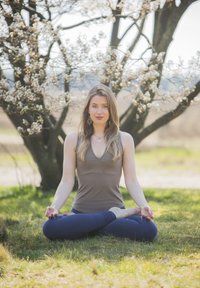 Nainen ruskeassa hihattomassa paidassa ja sinisissä legginsseissä istuu ristijaloissa ruohikolla, meditoimassa kukkivan puun alla kirkkaassa auringonvalossa.