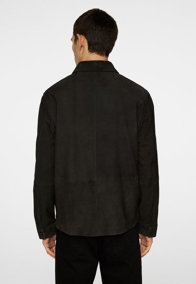 Man in een eenvoudige zwarte suede jas, gezien vanaf de achterkant, tegen een plain witte achtergrond.