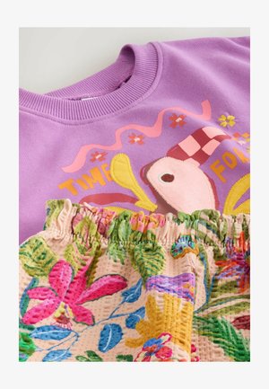 Sweat-shirt lilas avec un graphisme d'oiseau coloré et du texte, associé à un tissu à imprimé floral plissé sous l'encolure.